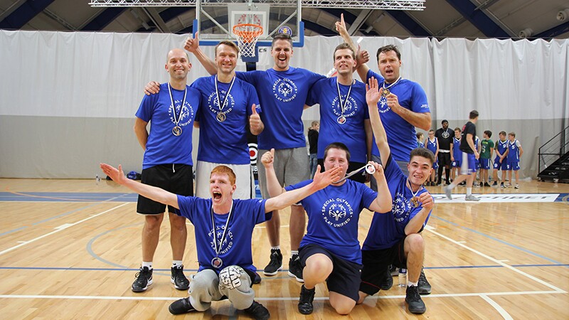 A Unified Basketball team standing in the middle of the gym posing for a group photo. There are two rows of players; the back row consists of 5 standing adult males and front row consists of three kneeling adults, two males on either side and one female in the center. 