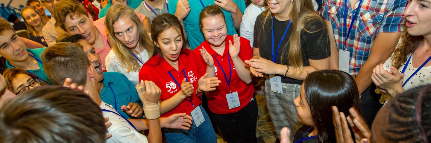 A large group of athletes and Special Olympics representatives stand in a circle clapping with joyful expressions on their faces.
