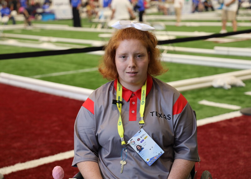 Emileigh Marsh sits in a wheelchair on an indoor turf competition field, wearing a gray and red Texas team shirt and a yellow credential lanyard. Bocce courts and event participants are visible in the background.