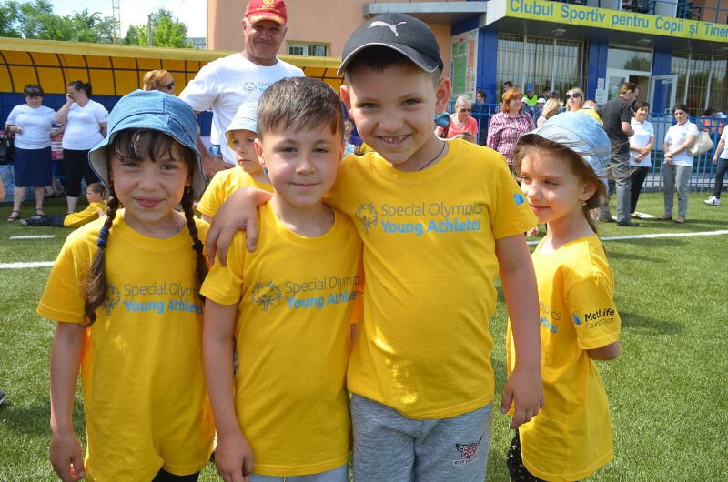 Three smiling young children in yellow Special Olympics Young Athletes t-shirts face the camera. They are standing on a grass pitch with a crowd behind them.