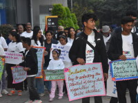 A large group of people walks together in a community inclusion awareness march on a city sidewalk, many wearing matching white event T-shirts and carrying colorful handmade signs promoting inclusion, diversity, equity, and belonging. In the foreground, participants hold posters with messages such as “Inclusion is the key to unlocking the potential of every child regardless of their abilities” and “Diversity is… Inclusion is… Belonging is…”. Adults and youth are participating side by side, with office buildings and landscaping visible in the background.