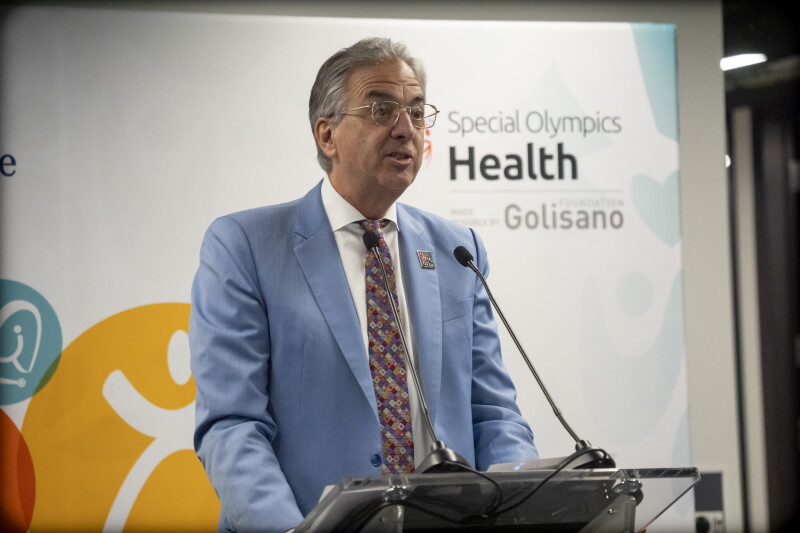A man in a blue blazer and patterned tie leans in to speak into a microphone at a podium. Behind him, a logo reading "Special Olympics Health Made Possible by the Golisano Foundation" is visible.