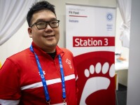A Special Olympics athlete standing in front of a Healthy Athletes Fit Feet banner. The banner is red, includes a graphic of a foot and highlights the third station of the screening. The athlete is wearing a red jacket and glasses, and is smiling for a photo. 