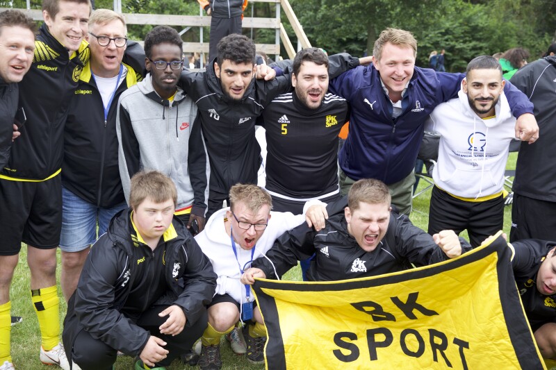 A group of men in sports gear huddle stand with their arms around each other in two tiers facing the camera. One holds a yellow banner saying 'BK Sport'.