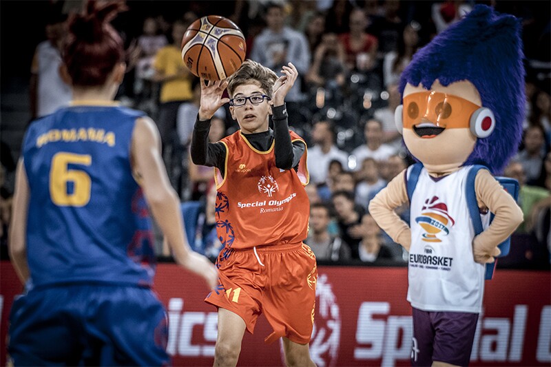 Young male Romanian player in an orange uniform catching the basketball. The 2017 Eurobasket mascot is in the middle ground. A girl is in the foreground wearing a blue uniform with the number 6 on her back. An audience is watching the action in the background.