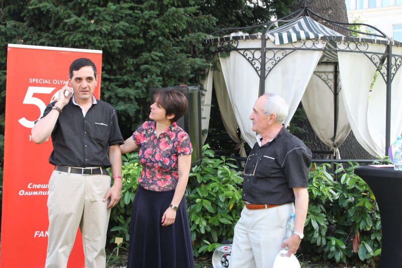 Two men and a woman stand in a garden setting in front of a Special Olympics 50 red pull-up banner.
