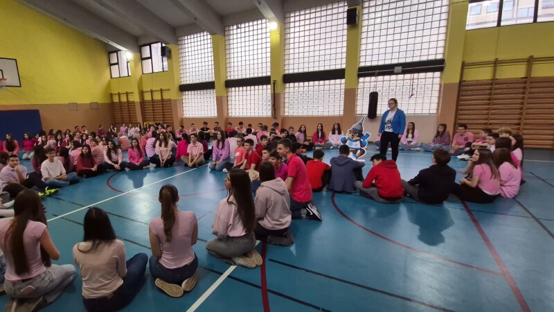 People sitting down in a gymnasium and one person standing and talking. 