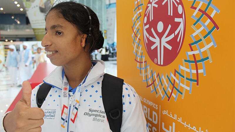 A young woman standing in front of the Athletes Chill Out sign at the 2019 World Games giving a thumbs up and showing off her new hearing aid.