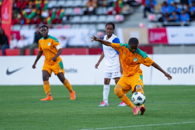 Three players, two from a team in orange and turquoise, one kicking the ball, and the opposing in white and blue.