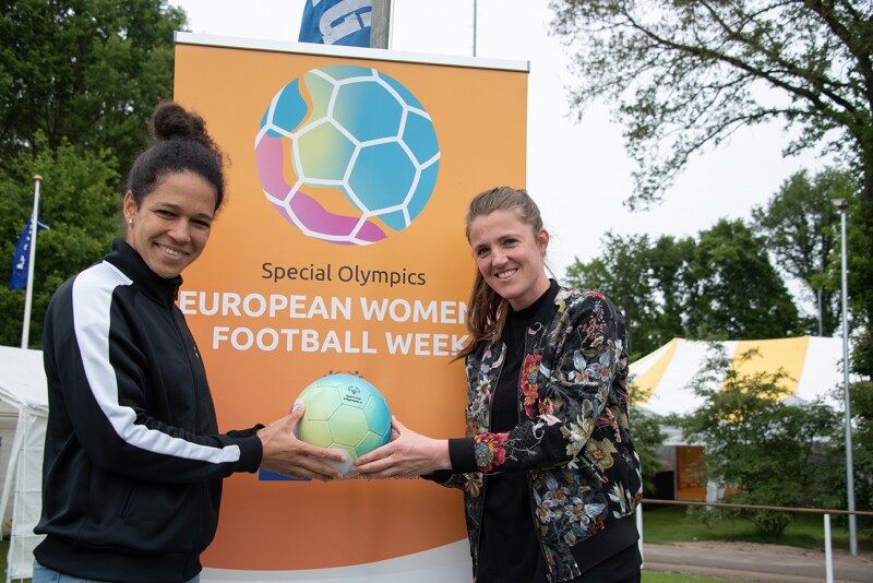 Two women stand holding a ball in front of an orange Special Olympics European Women's Football Week banner.