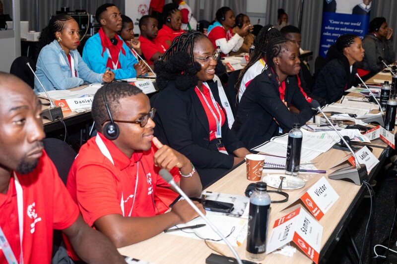 Youth leaders sit along two summit rows. Their desks have microphones and name tags. They are smiling.