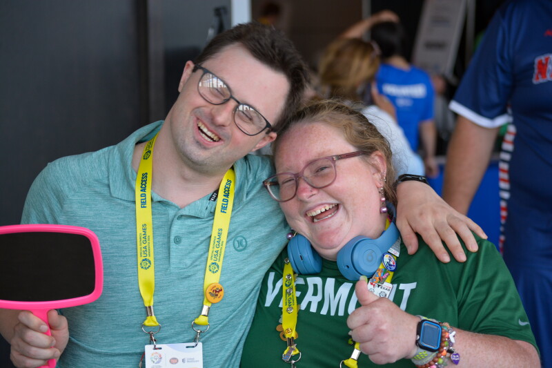 Two people wearing glasses stand smiling with their arms wrapped around each other. The man on the left holds a mirror and the woman on the right does a thumbs up motion. 