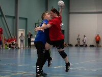 A woman wearing a red jersey jumping, holding a ball  colliding with a man on the opposing team, wearing a blue jersey