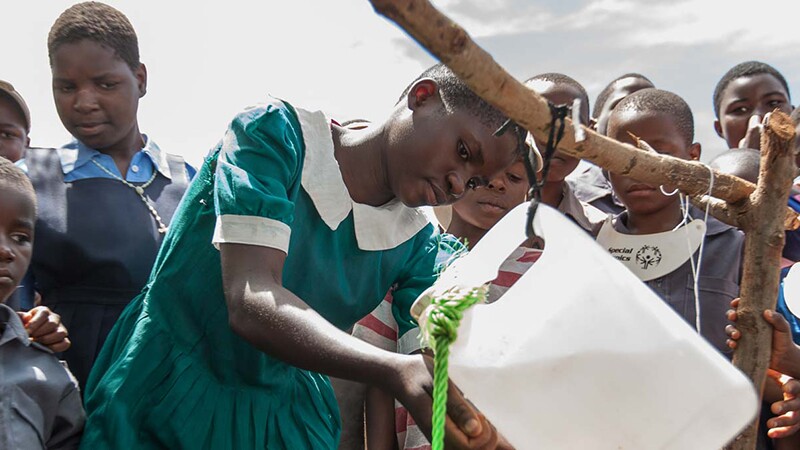 A young girl is washing her hands from a water jug and young children are around watching her.