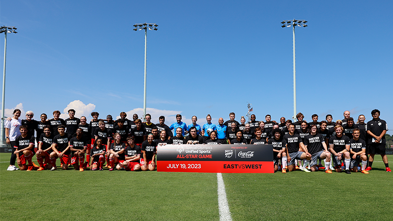 A group of Special Olympics soccer players, coaches and officials pose for a pre-game photo on the soccer field. They're standing behind a sign that says "Special Olympics Olympics Unified Sports All-Star Game". 