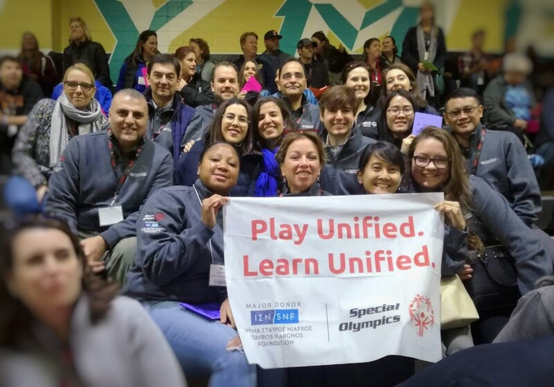 A group of people sitting in a sports stand face the camera holding up a white banner with the words 'Play Unified. Learn Unified' and the logos of Special Olympics and the Stavros Niarchos Foundation.