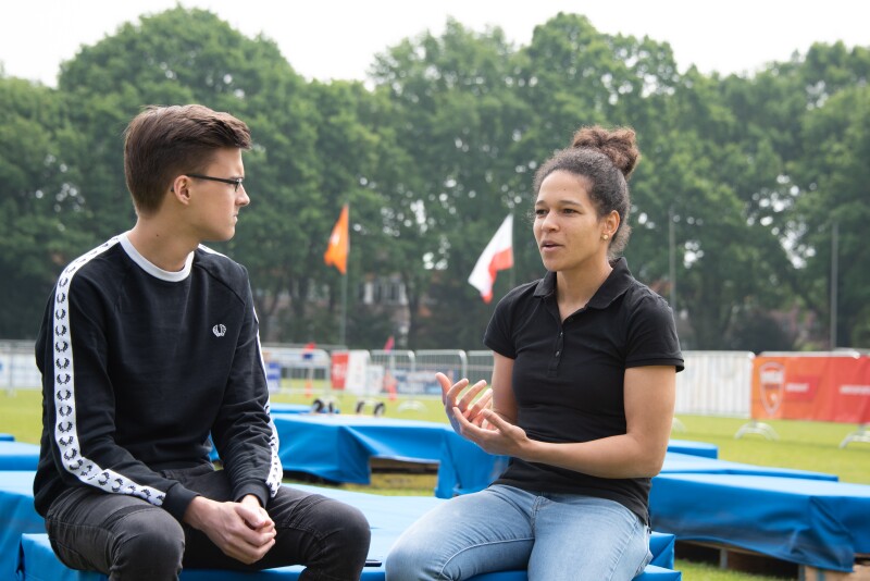 A man and women sit at the side of a football pitch in conversation. Special Olympics flag and hoarding in the background.