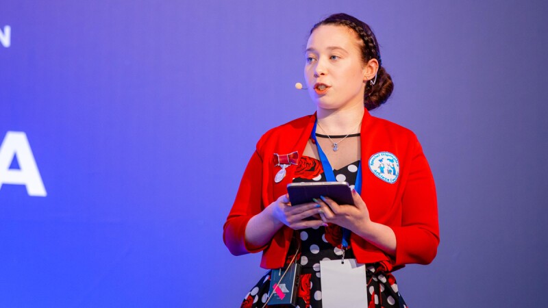 Woman wearing red jacket and a dress speaks on the stage