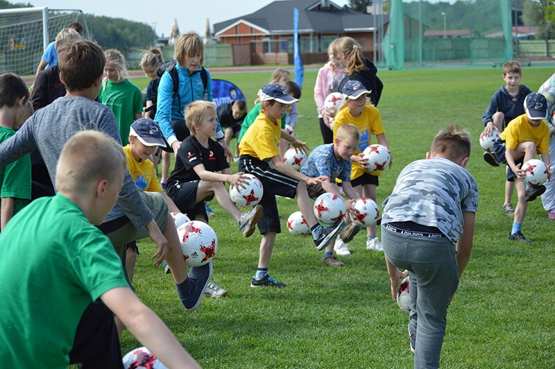 Young athletes on the field balancing a football on their foot for coordination practice. Adults are assisting and watching their progress.