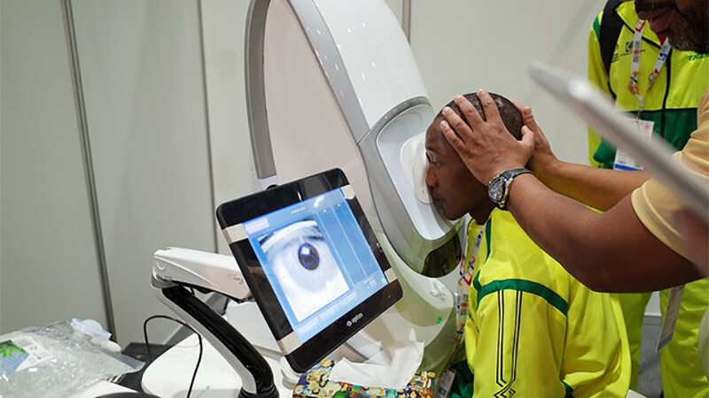 Mohammed Mubarak receives an eye screening at Opening Eyes; his face is being pressed against a machine by a volunteer and his eye is being examined and photographed by a machine.