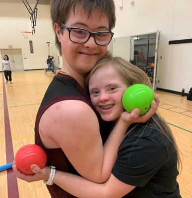 Two athletes share a moment of friendship during a bocce activity, holding bocce balls on an indoor gymnasium court.