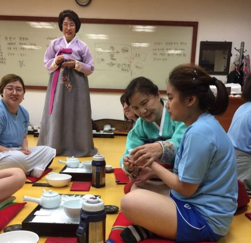 Small group in a room with a white board in the background. Woman standing is wearing kimono, athletes and partners are sitting, speaking, and drinking tea.