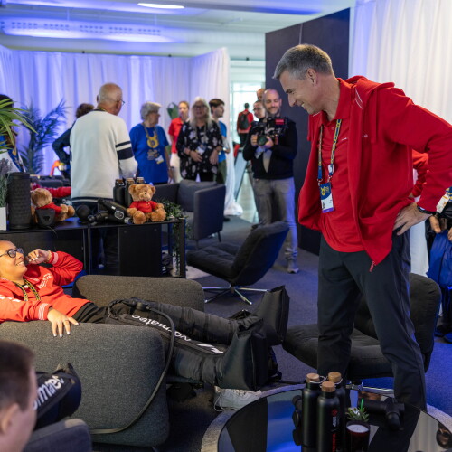 In a busy room full of people, a man in a recliner chair smiles up at a man standing near him.  