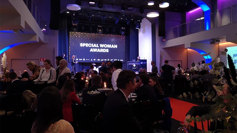 Special Woman Awards illuminated on a projection screen in the front of a hall with people sitting in the foreground. 