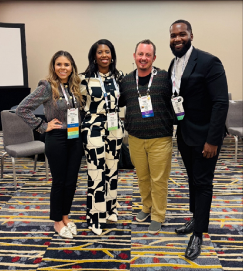 2 women and 2 men smile in a group picture. Behind them, there is a round circle of chairs from the coffee meeting that they hosted. 