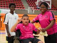 A smiling woman embraces her son, who is on a wheelchair, while her other son stands behind them, also smiling.