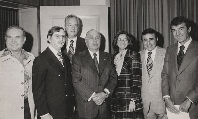 1969-black-and-white-photo-of-10-men-and-women-posing-in-suits-and-dresses-in-front-of-drapery.jpg