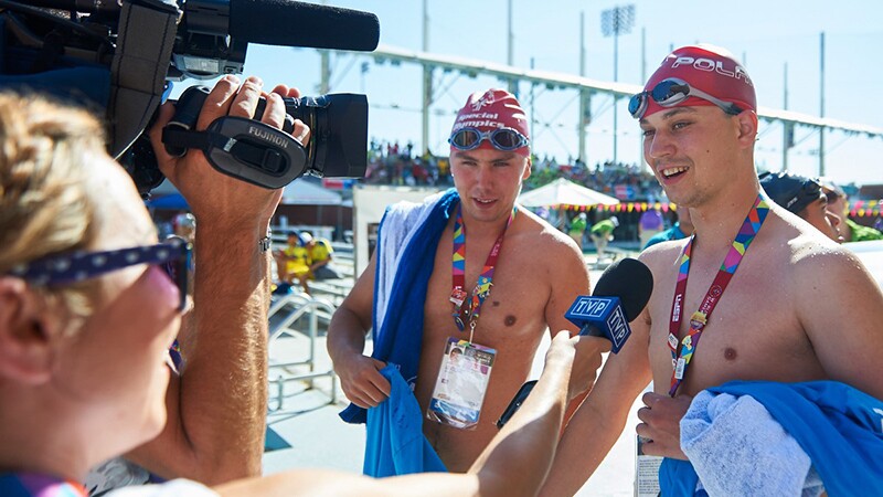 Two men in swimming caps holding towels face a camera and speak to a woman holding a microphone.