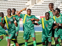 A group of footballers in green uniforms joyfully celebrating on a soccer field, raising their arms and smiling after a victorious goal.