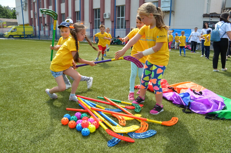 Children in yellow t-shirts holding colourful plastic hockey sticks with other colourful equipment and balls laid out on the grass beside them. 