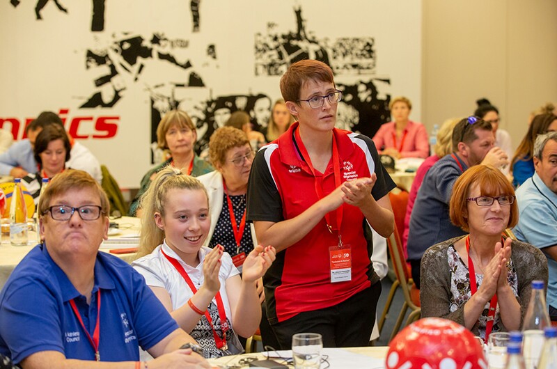 Athlete leaders Micheline Van Hees, Kiera Byland and Johanna Pramstaller with Jacqui Byland at the Special Olympics Europe Eurasia Leadership Conference 2018. One athlete is standing and speaking in the crowd and listeners are applauding.