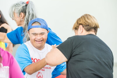 Two participants dance together during a Special Olympics Dance Olympics event. One participant wearing a Special Olympics shirt and blue cap holds hands with another participant as they take part in an inclusive dance activity, while others dance nearby.