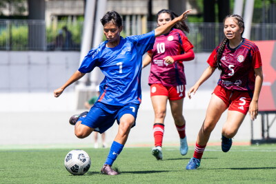 A male player prepares to kick the ball while two women from the opposing team come up behind him. 