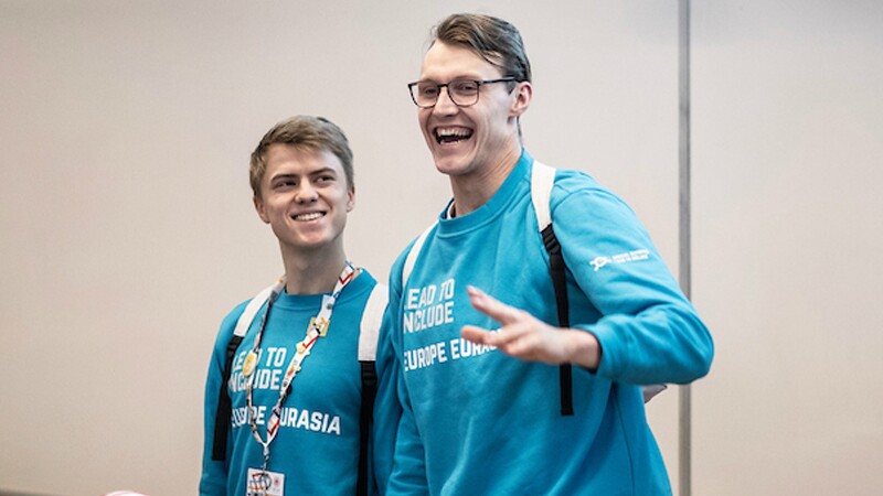 Two young man standing side by side, the young man on the right is waving to someone off camera.