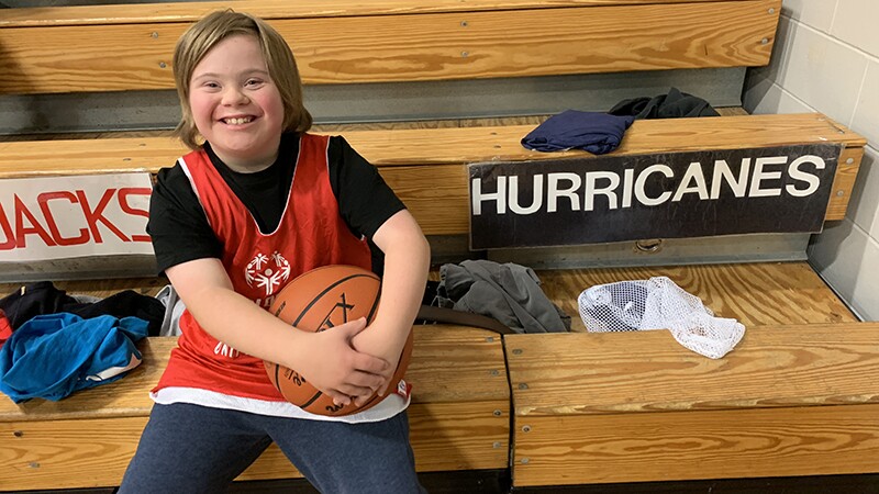 Boy smiling sitting on bleachers holding a basketball wearing a red Special Olympics tank top with a team sign, "Hurricanes" in the background.