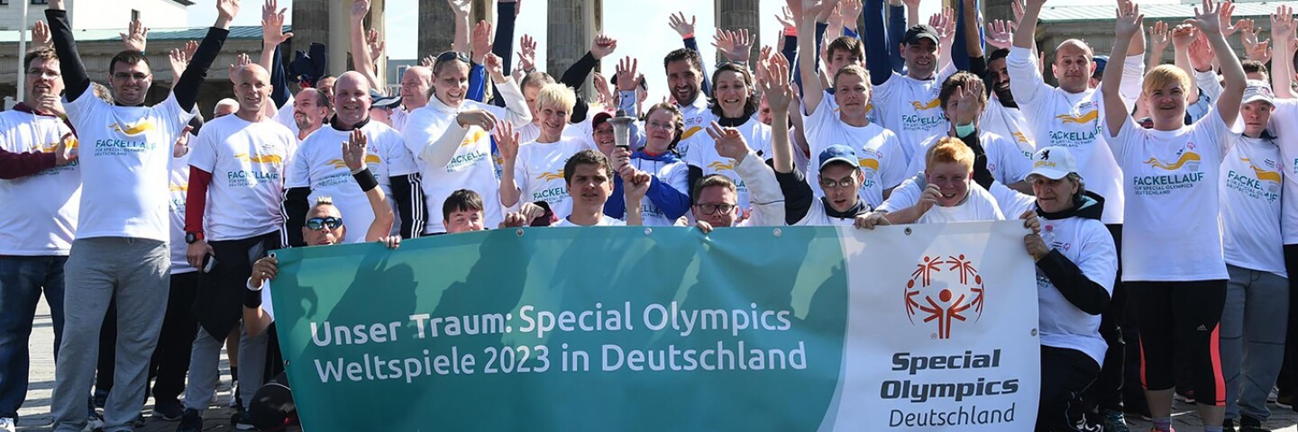 a large group of athletes and representatives cheering with their hands up and some are holding a banner in front of them.