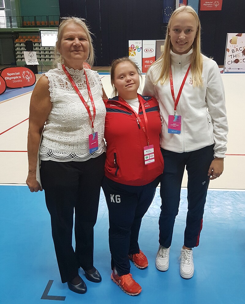 Three women – all with lanyards around their necks and two of whom are in tracksuits – stand side by side facing the camera and smiling. In the background is a gym floor, seating and banners.