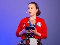 Woman wearing red jacket and a dress speaks on the stage