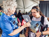 An older woman in a blue shirt assists a younger woman by showing her a mirror at a crowded indoor event.