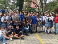 A large group of students gathers outdoors on a school campus holding badminton rackets. Participants pose together on a paved court near a badminton net, with trees and fencing in the background during a youth sports and inclusion activity.