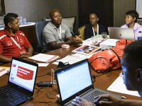 Six people sit around a table with computers, folders, pens and paper. Jimmy Masina addresses them from his seat.