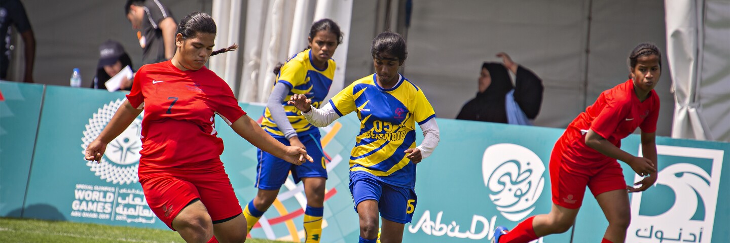 Four young women on the field playing football, two from each team with spectators in the background.