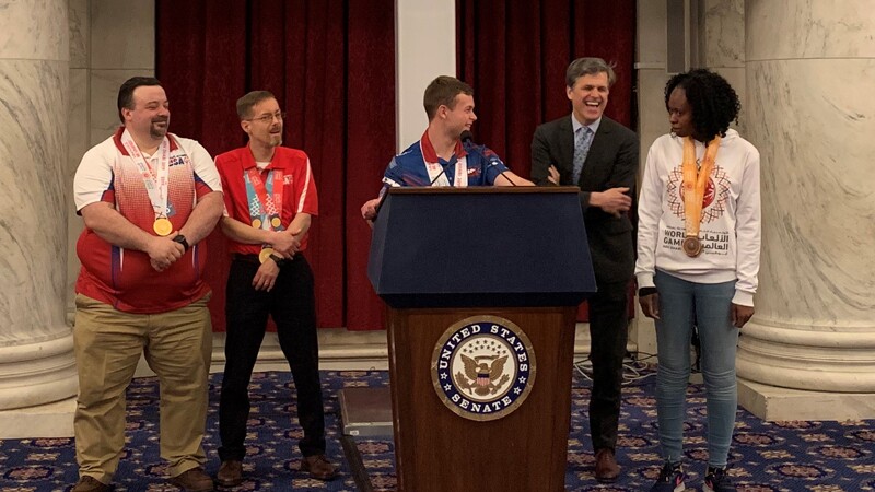 Five people stand at a podium at the U.S. Senate. They are framed by the U.S. flag on the left side and by the Senate flag on the right side.