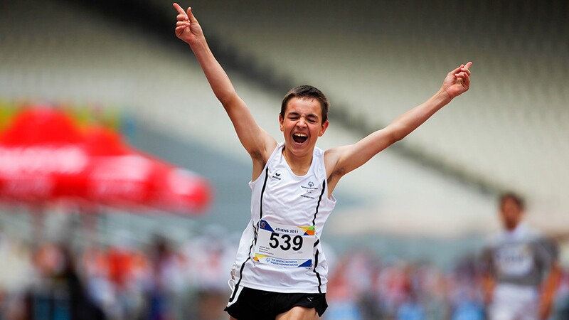 Young man in a white tank top with two thin black stripes, on either side and black running shorts; he's finishing a race with his hands up and index fingers pointing up (#1) with an expression of success/victory; his bib number reads 539