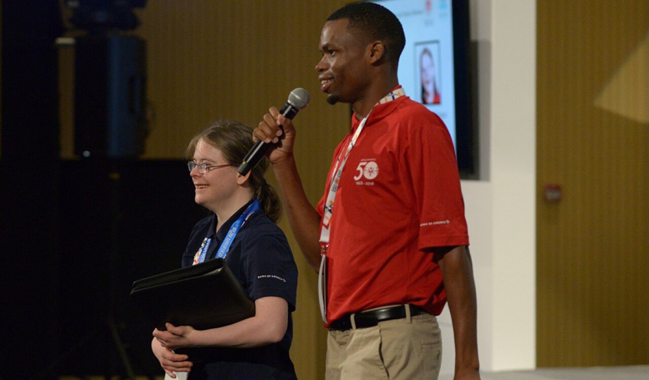 Two young adults standing on stage speaking into a microphone.