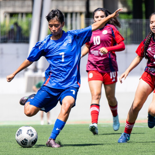 A male player prepares to kick the ball while two women from the opposing team come up behind him. 